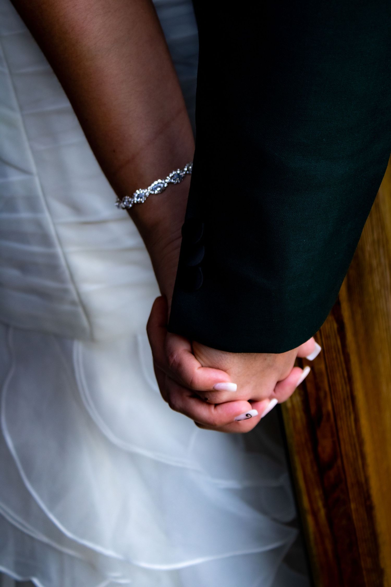 Close-up of intertwined hands with delicate crystal bracelet against wedding attire.