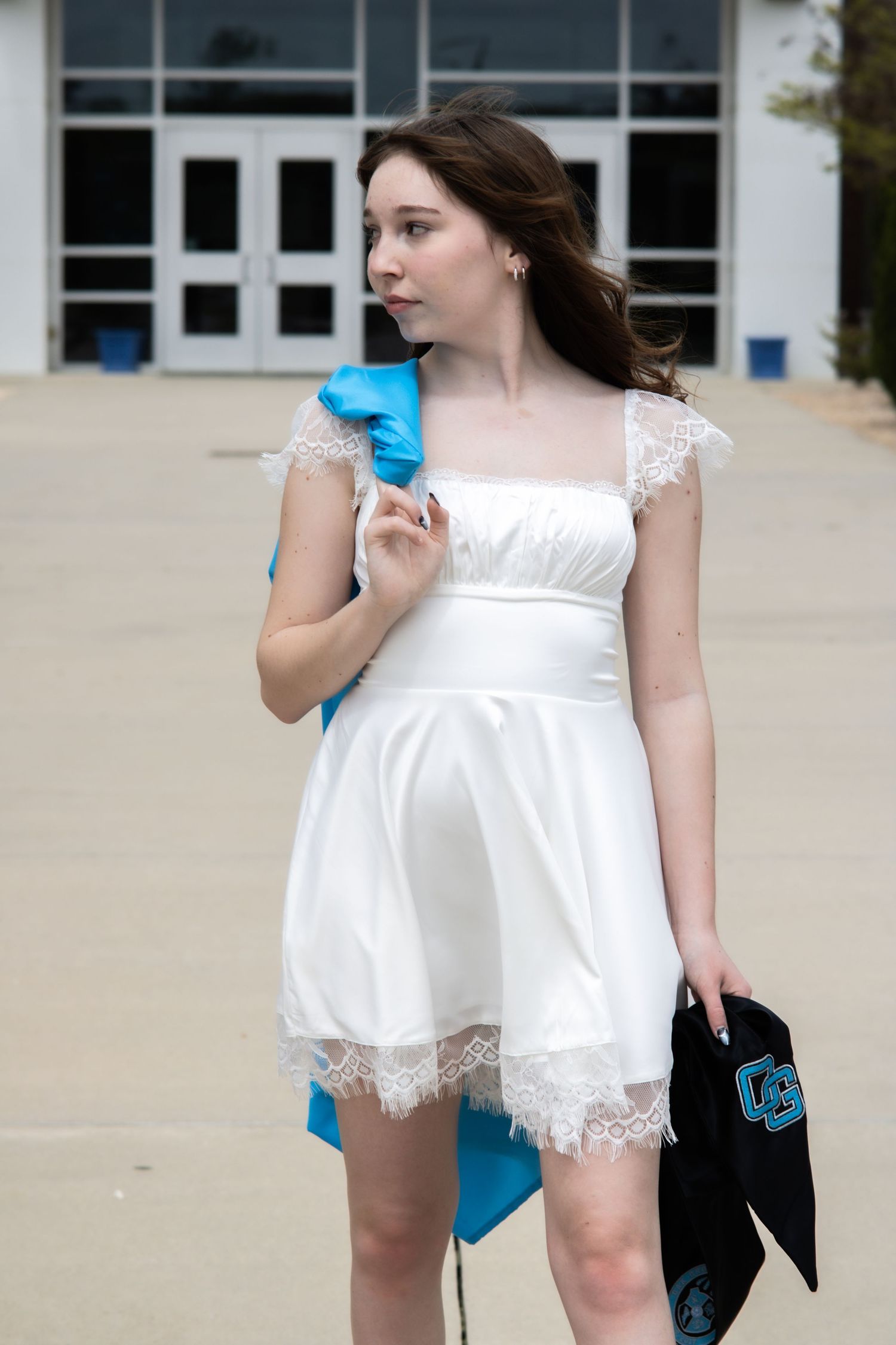 Graduate in white lace dress holds blue graduation gown outside academic building.