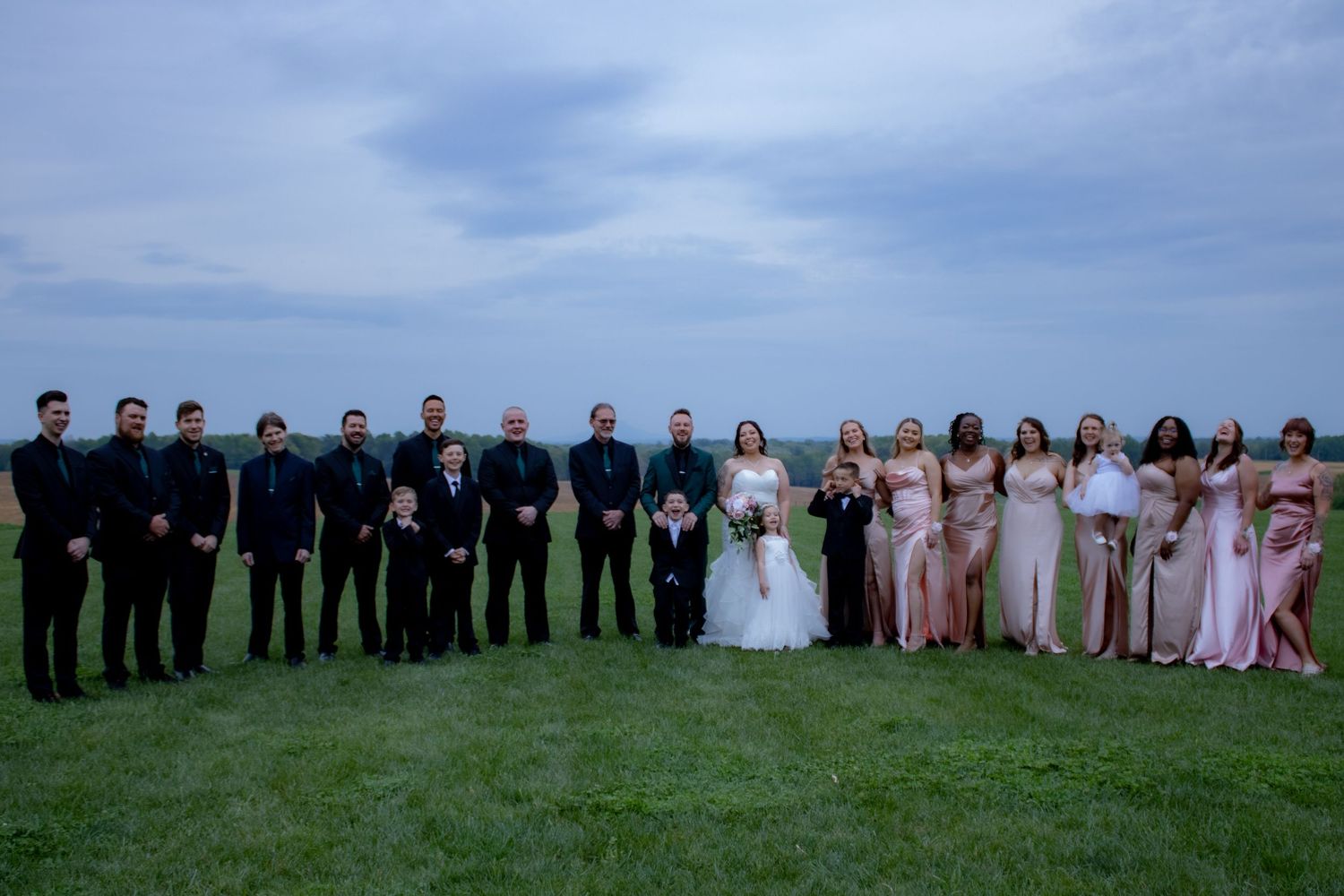 Wedding party lineup at dusk with groomsmen in black and bridesmaids in pink dresses on a grassy hill.