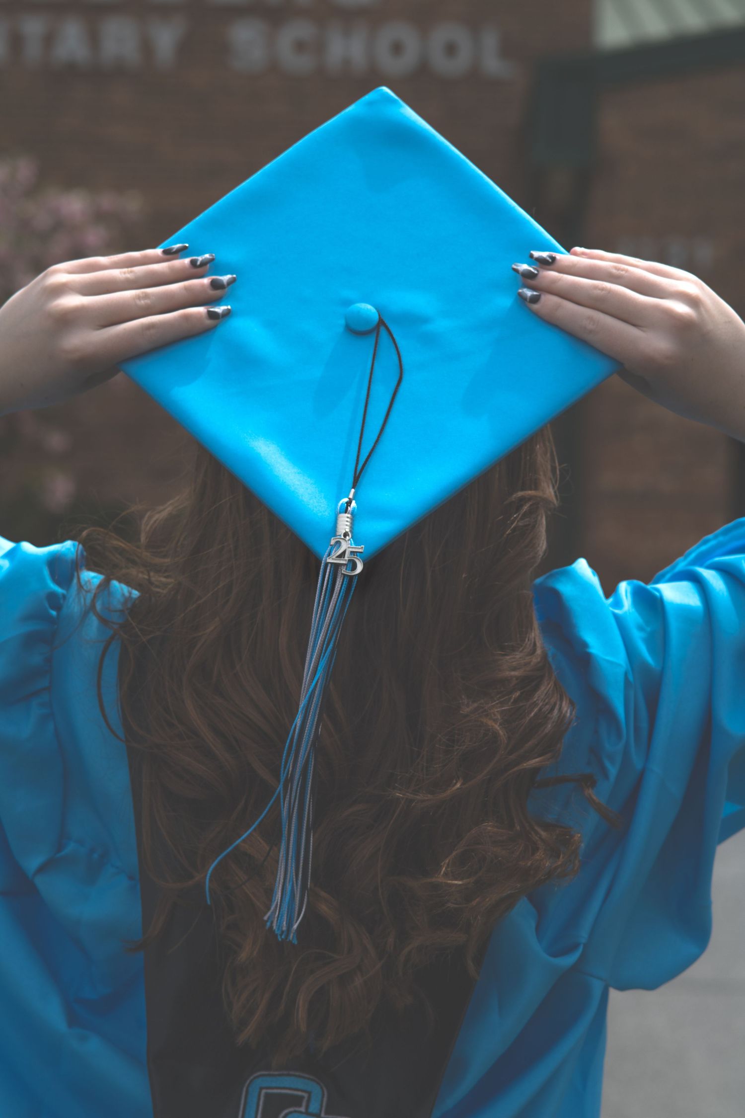 Close-up of hands holding bright blue graduation cap with tassel against school building.
