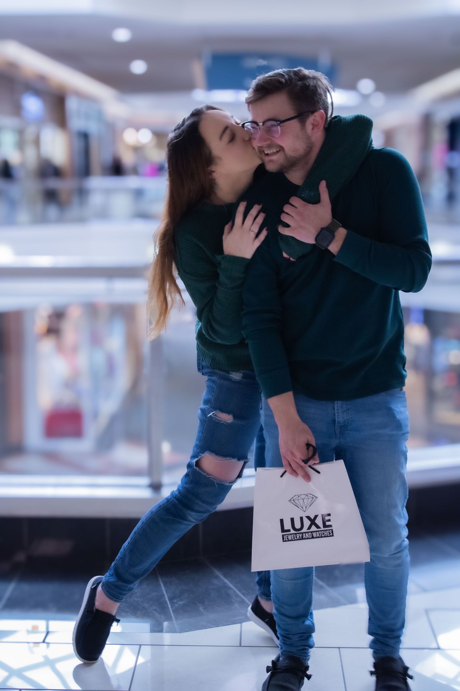A romantic couple leans against a glass railing in a shopping center with a 'LUXE' shopping bag.