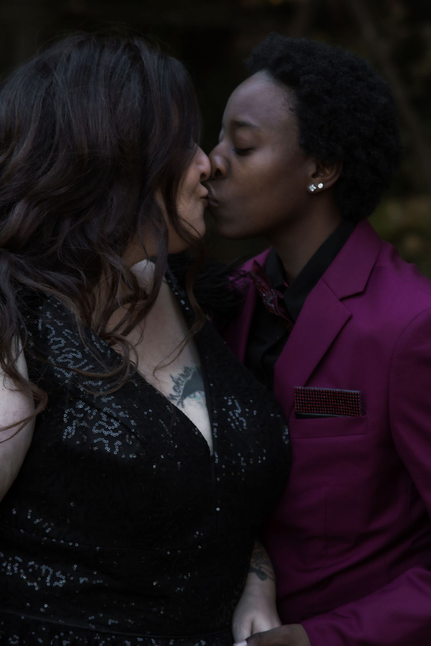 A couple shares a romantic kiss, one wearing a magenta blazer and the other in a sparkly black dress.