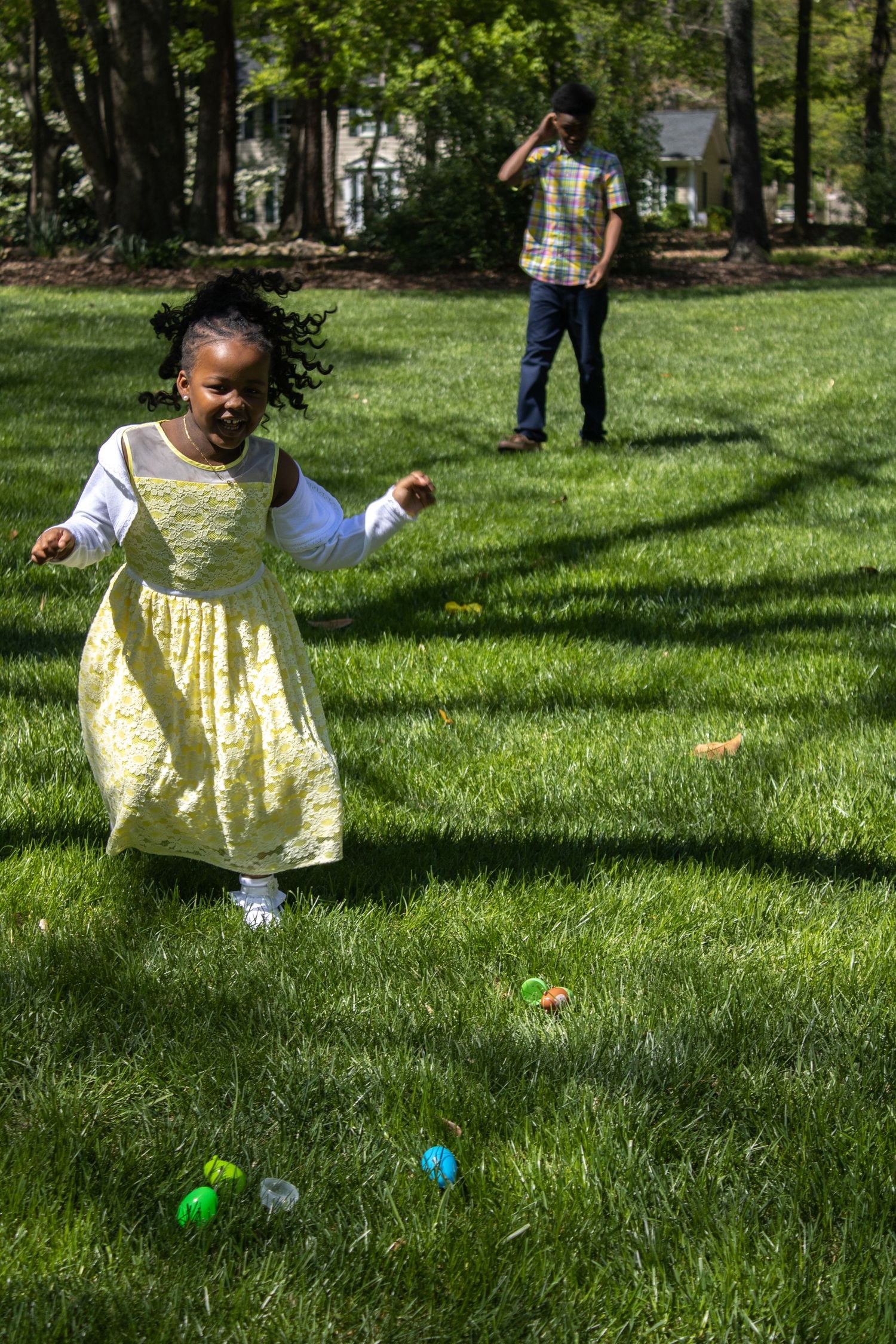 A young child in a yellow dress hunts for Easter eggs on a grassy lawn while someone watches from behind.