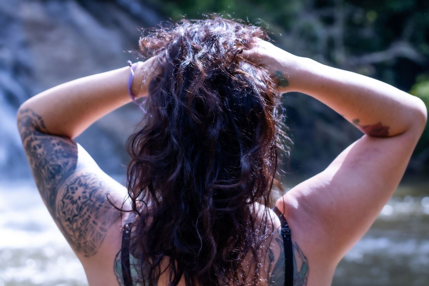 Back view of person with long dark hair and visible arm tattoos outdoors.