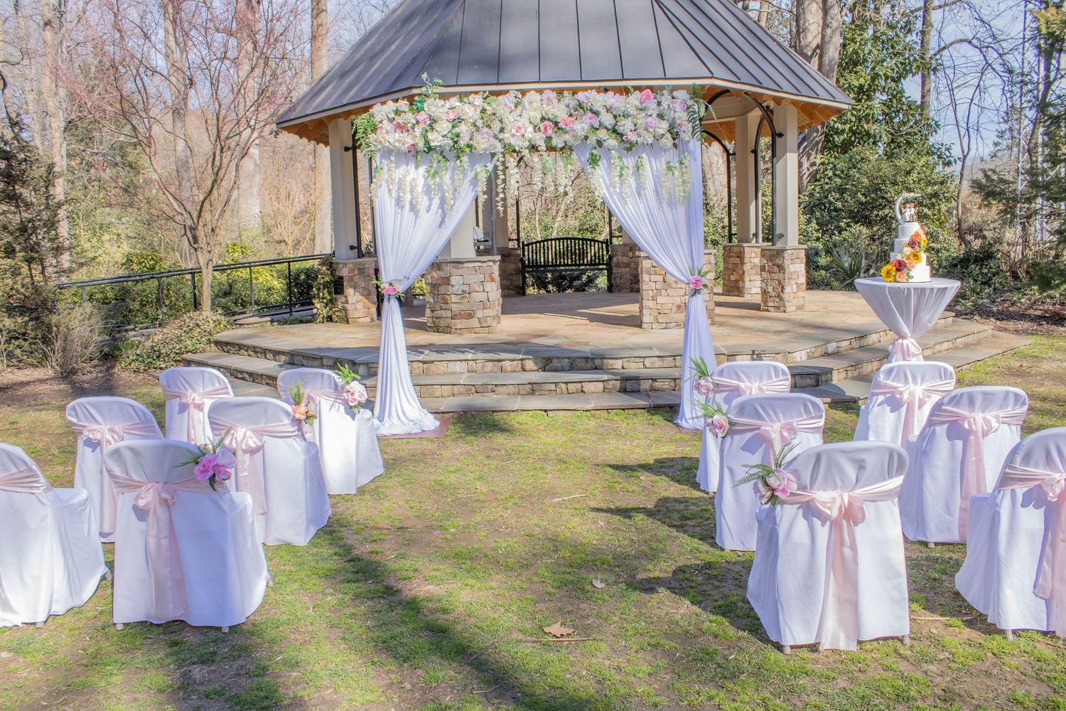 Outdoor wedding ceremony setup with white chairs and gazebo decorated with floral arrangements.