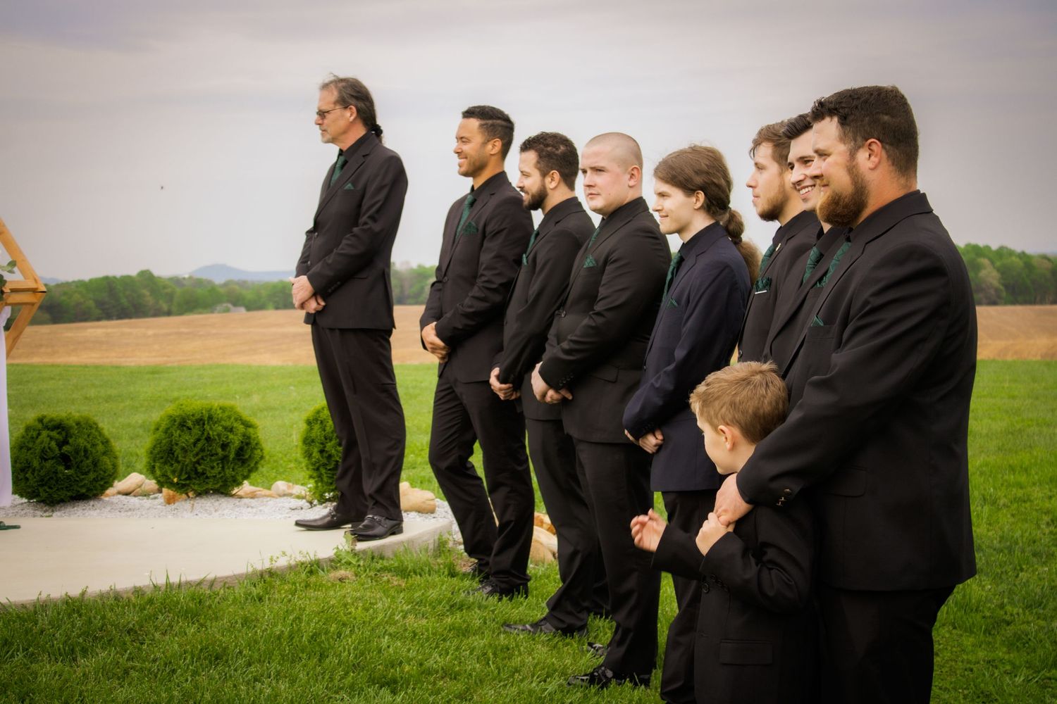 Wedding party members in black attire standing in a line outdoors against a rural landscape.