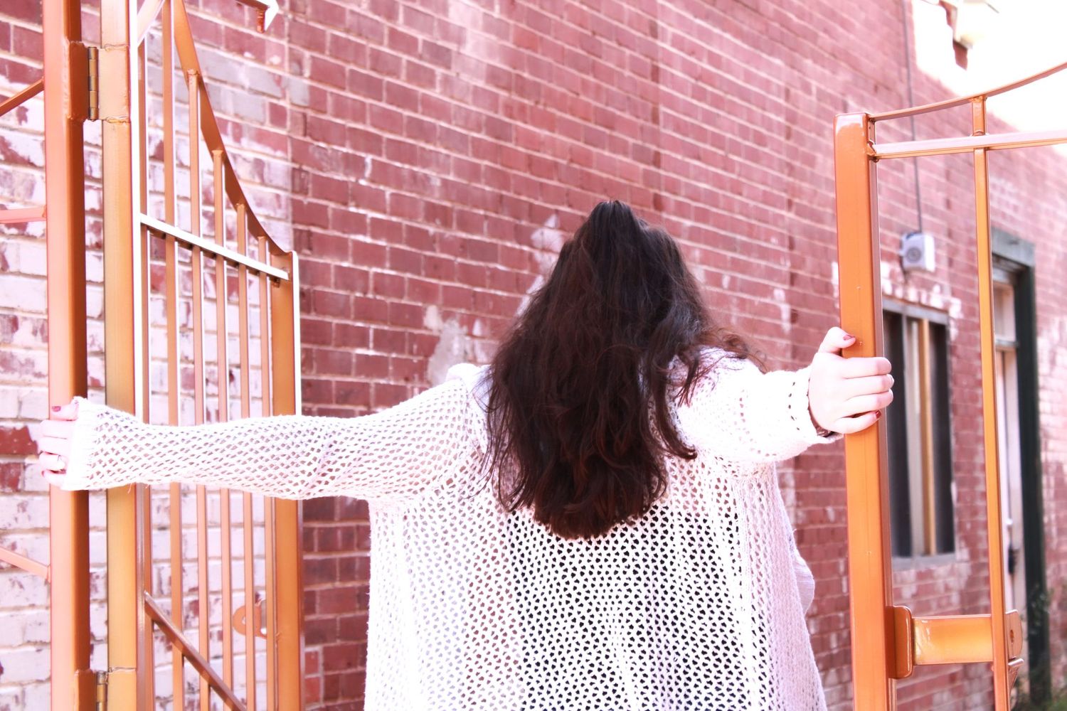 Person with long dark hair wearing white knit sweater photographed from behind against brick wall.