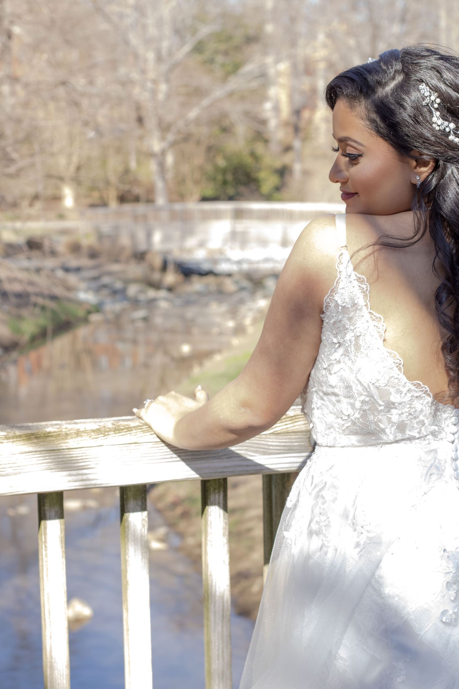 Bride in elegant lace dress gazes over railing with sunlit nature backdrop.