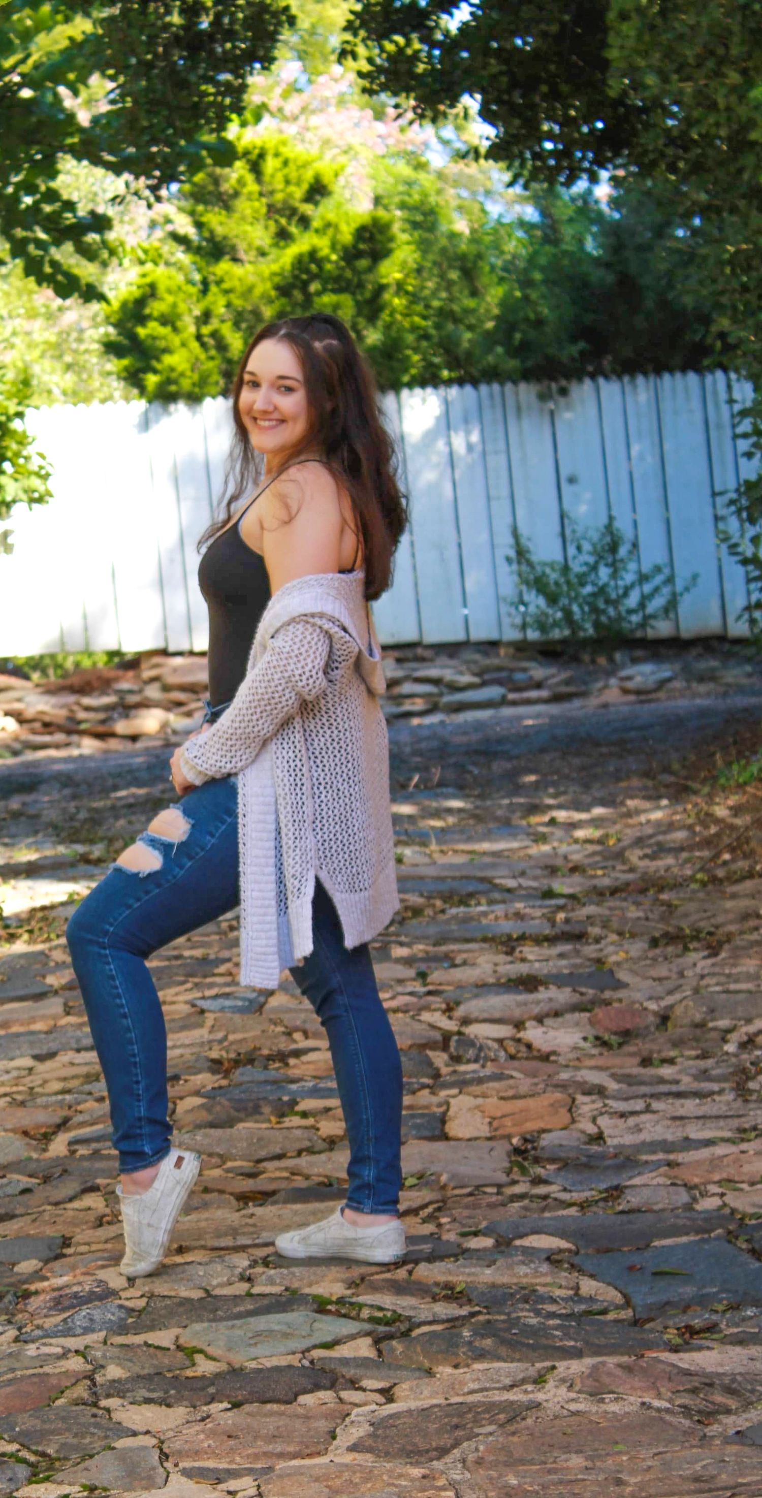 Woman in casual outfit poses on stone pathway with white fence in background.