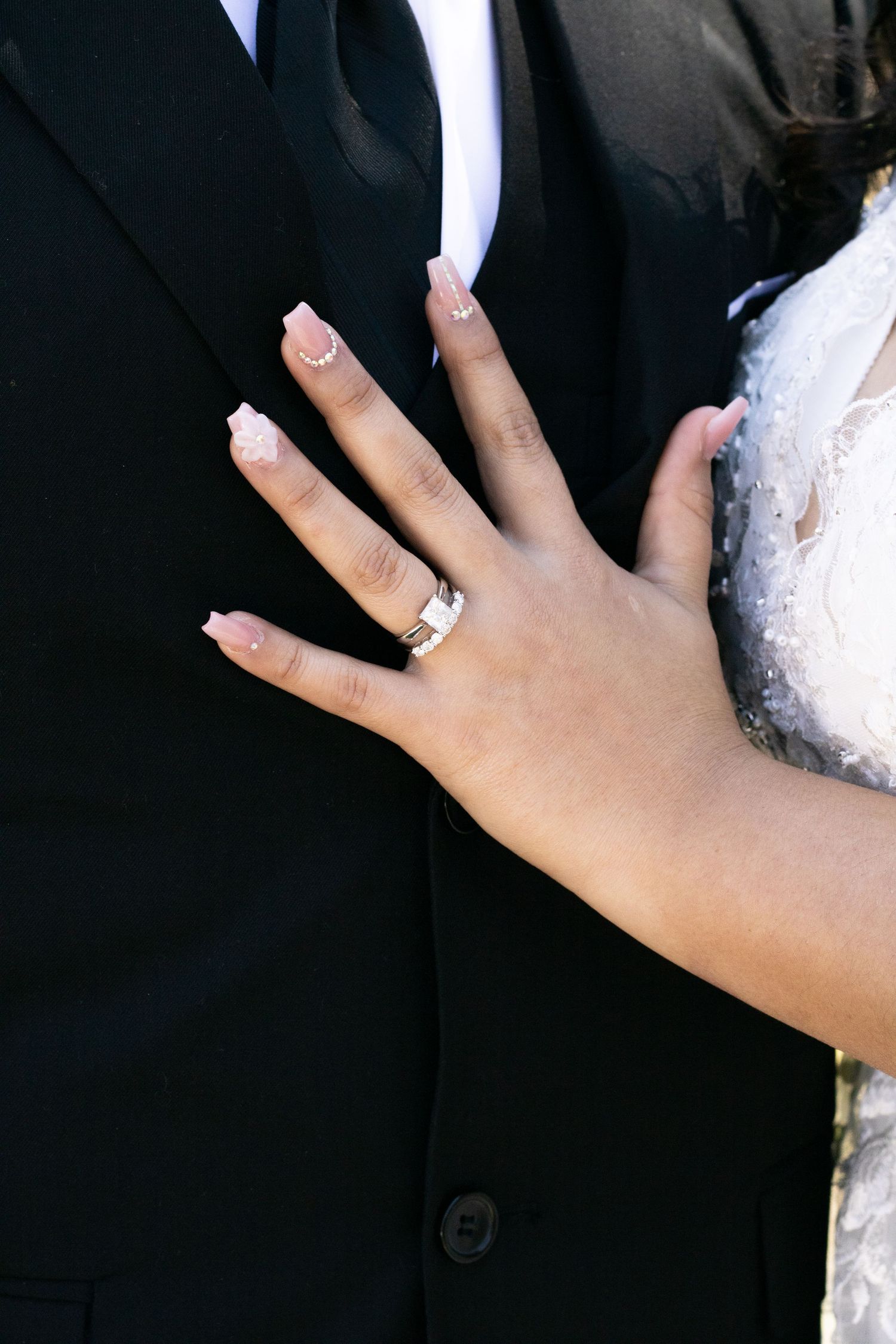 Close-up of a bride's hand with elegant manicure showing off wedding rings against groom's black suit.