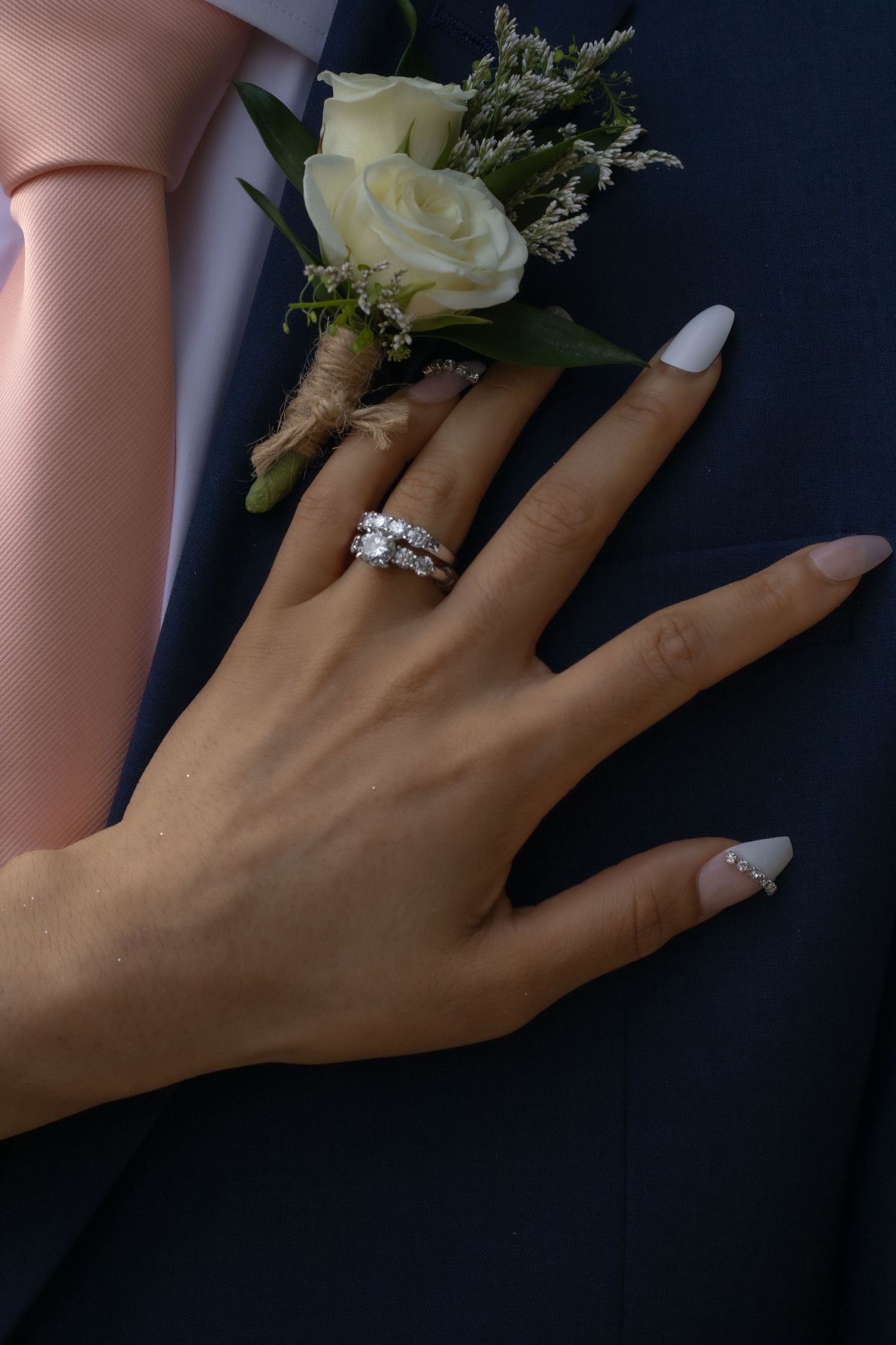 Wedding ring details with delicate white rose boutonnieres against dark background.