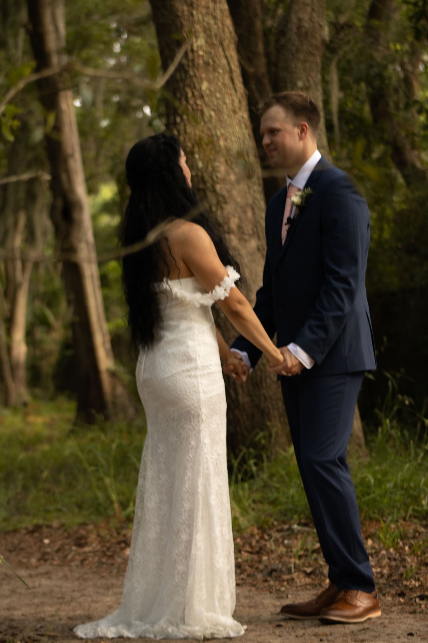 Bride in a fitted lace gown holds hands with groom in navy suit during their forest wedding ceremony.