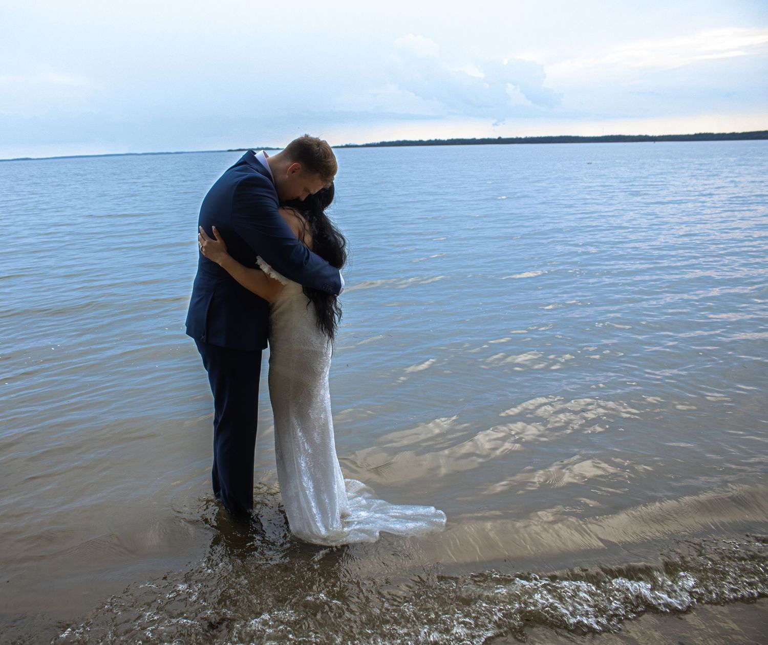 Couple in wedding attire embrace at the water's edge during sunset.