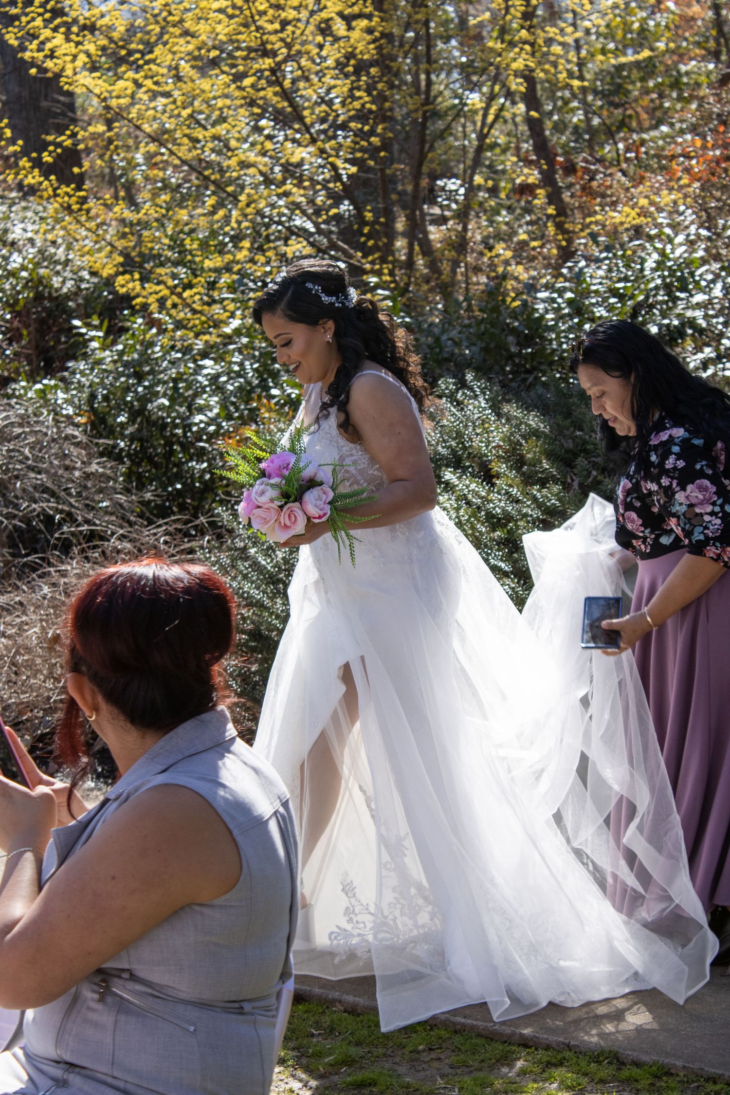 Wedding party helps adjust flowing white bridal gown outdoors in autumn setting.