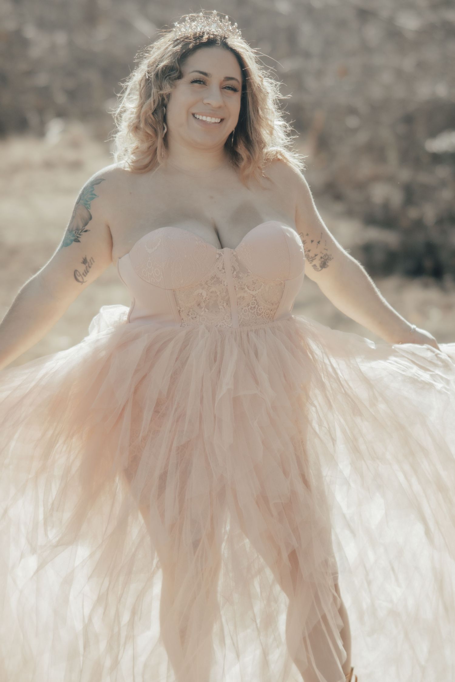 Woman in pink tulle gown poses outdoors in soft natural lighting wearing a crown.