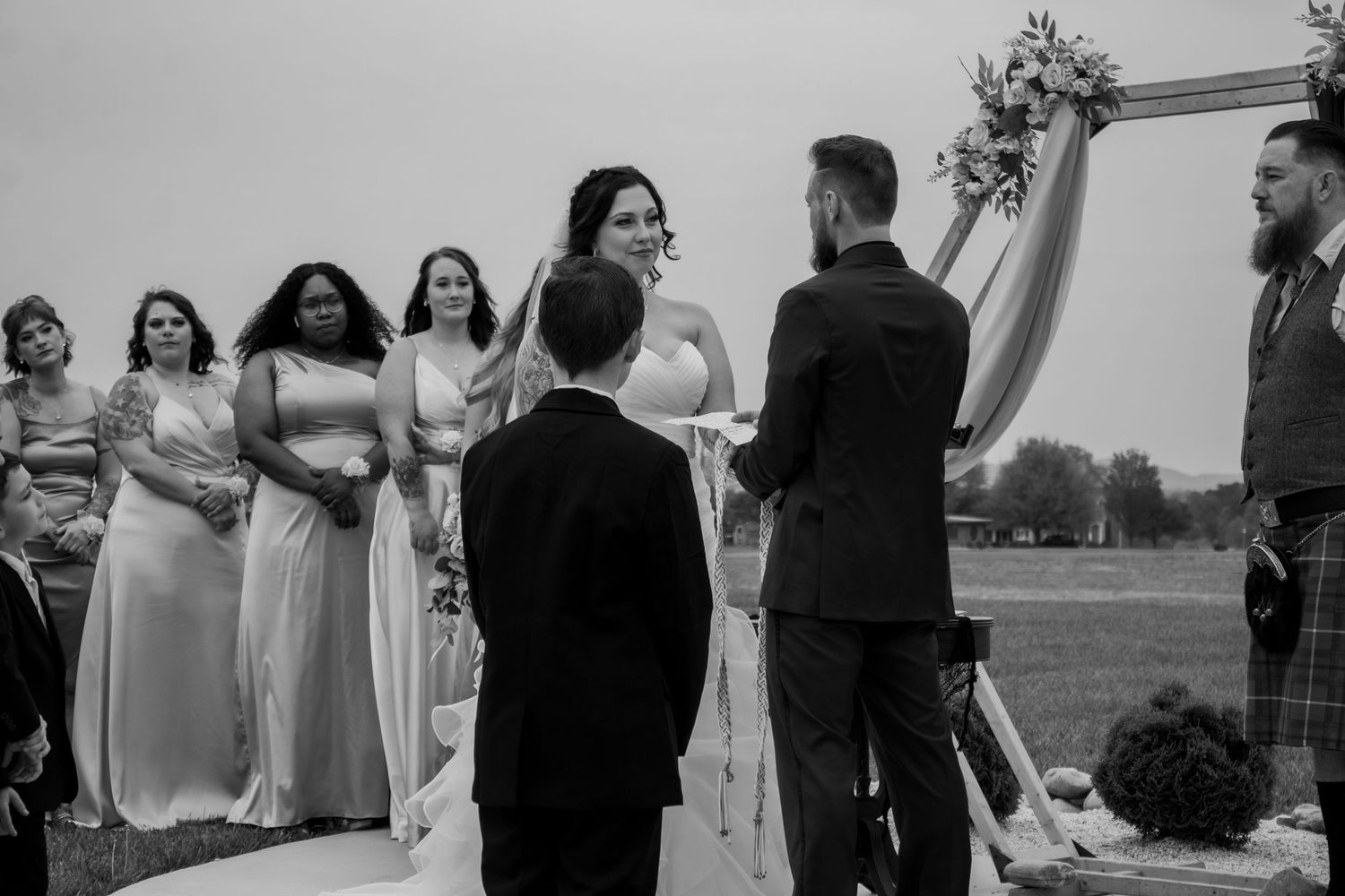 Outdoor wedding ceremony under a floral arch with attendants in light colored dresses.