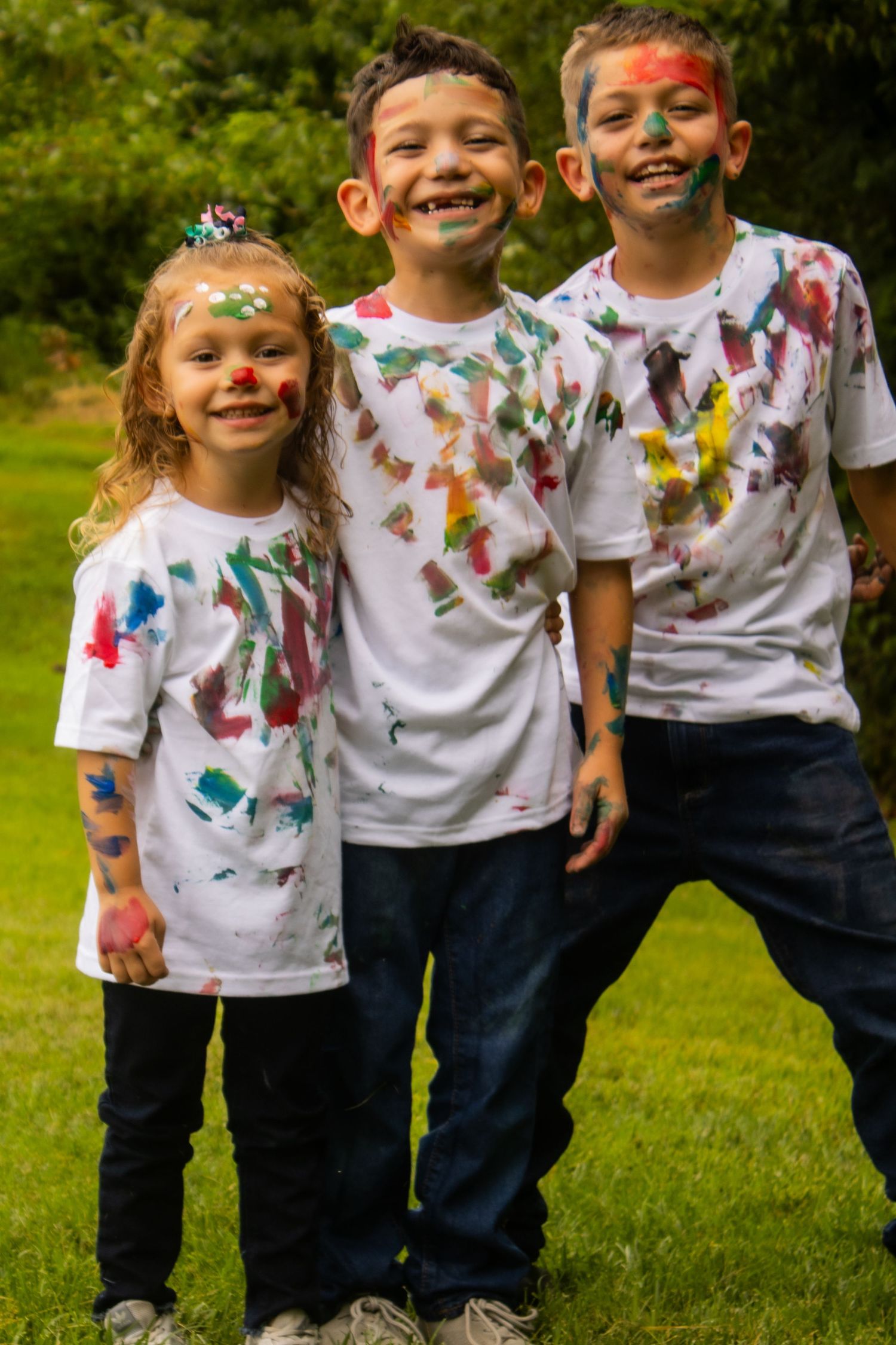 Three children wearing paint-splattered white t-shirts smile brightly at camera.