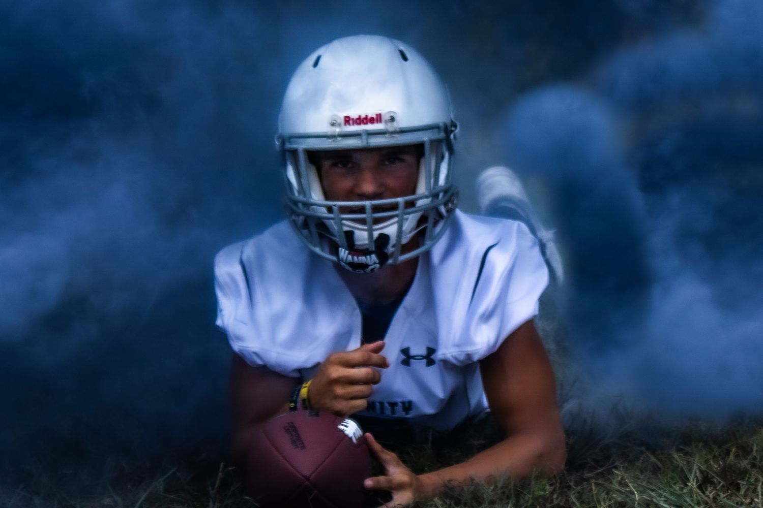 A football player in white uniform and silver helmet crouches with the ball in atmospheric lighting.