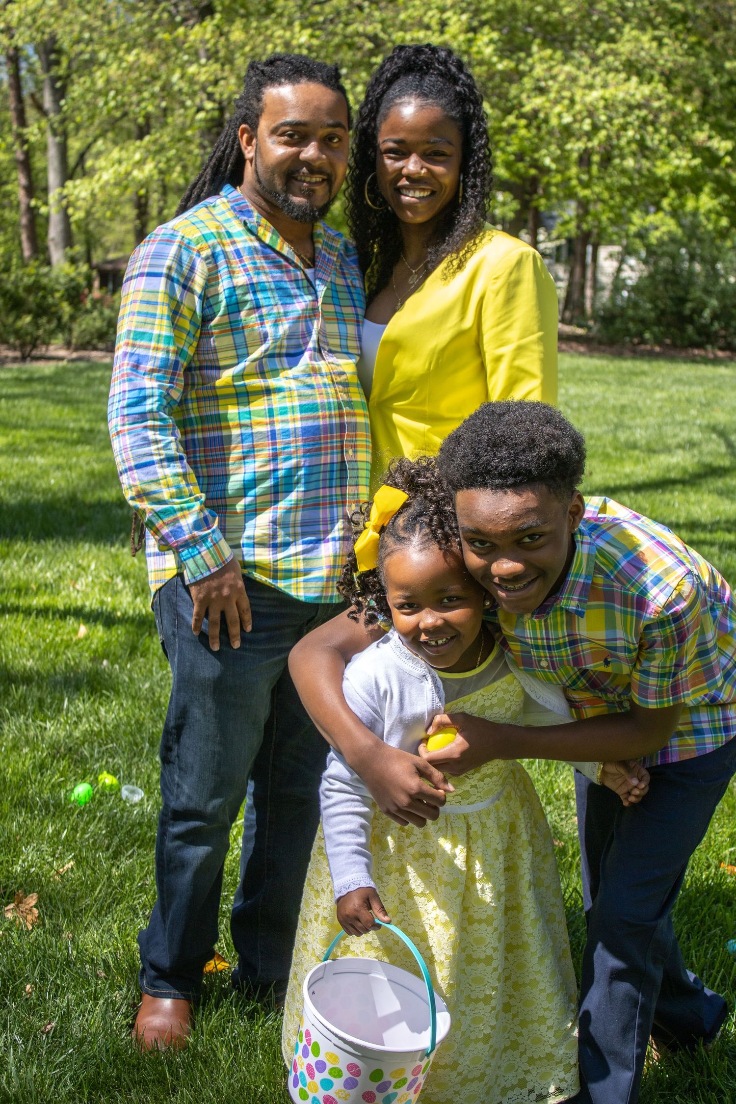 A family in coordinating plaid and yellow outfits poses together on a sunny lawn with Easter eggs.