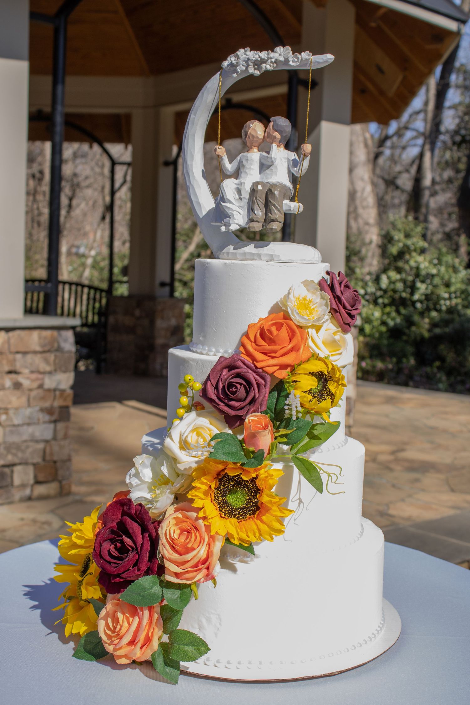 Four-tier white wedding cake decorated with colorful roses and sunflowers, topped with a romantic cake topper.