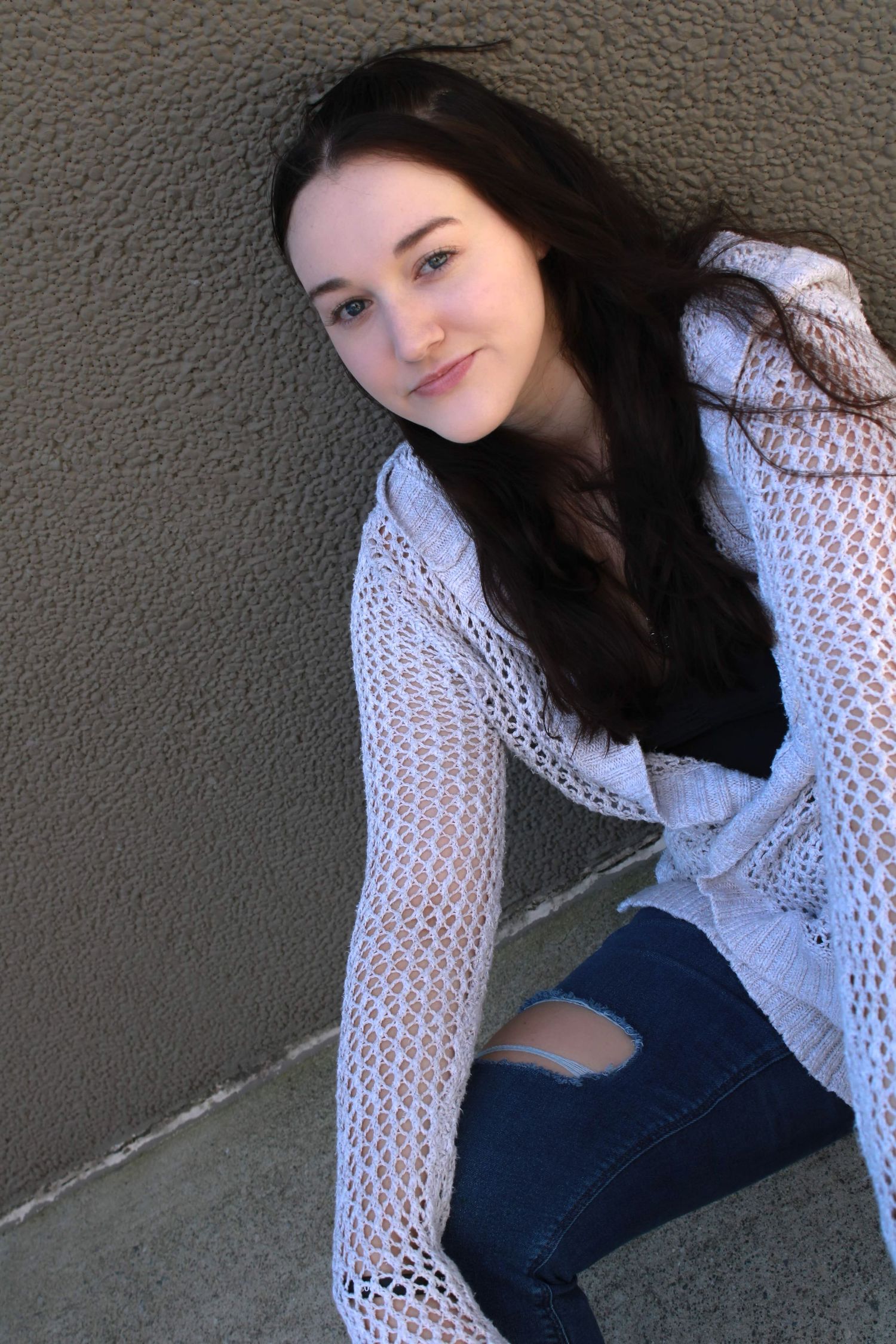 Young woman in white lace sweater and ripped jeans poses against gray wall.