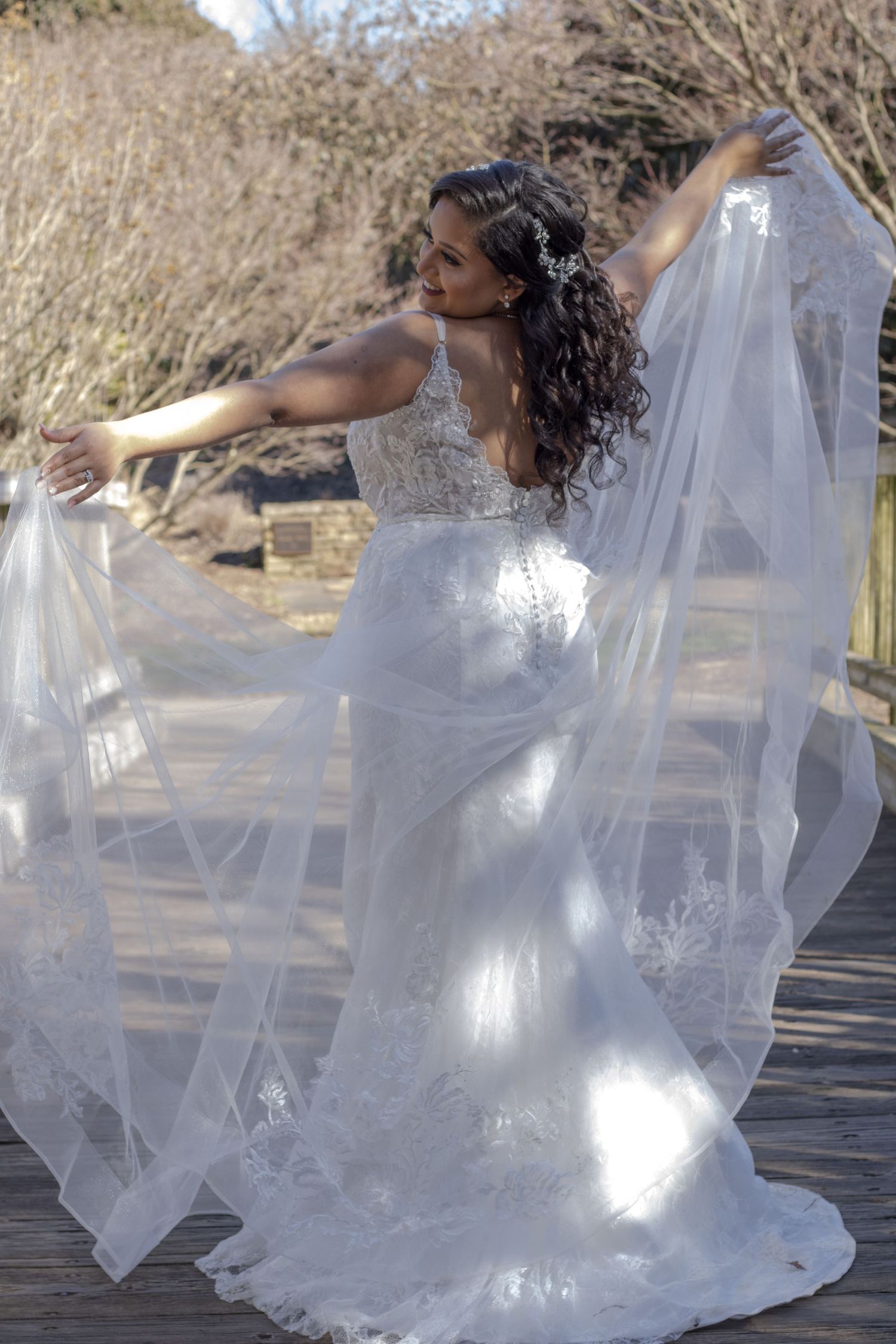 Bride in lace dress poses gracefully with flowing veil on wooden deck.