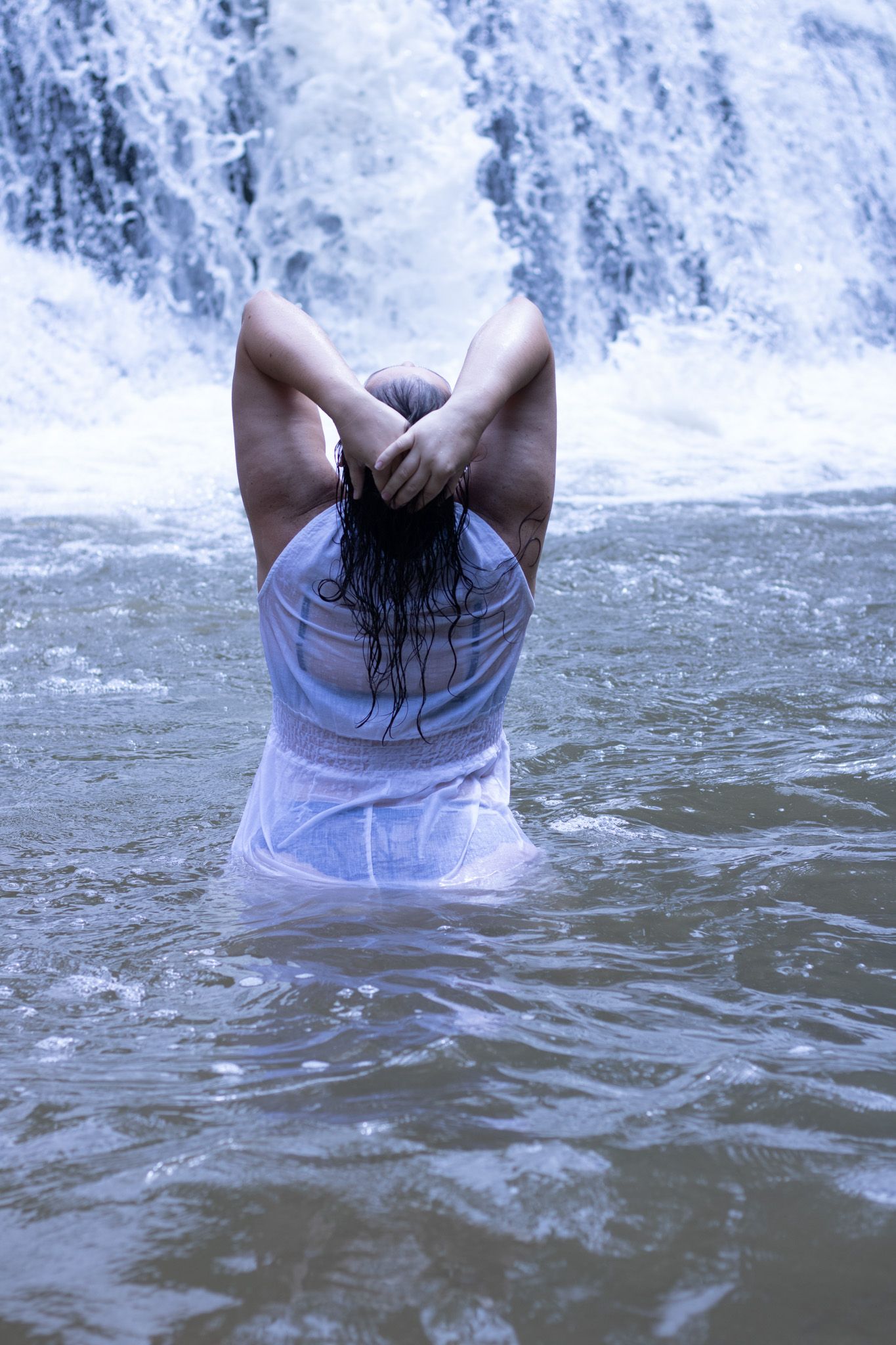 Person enjoying waterfall spray while standing in flowing water wearing wet white clothing.