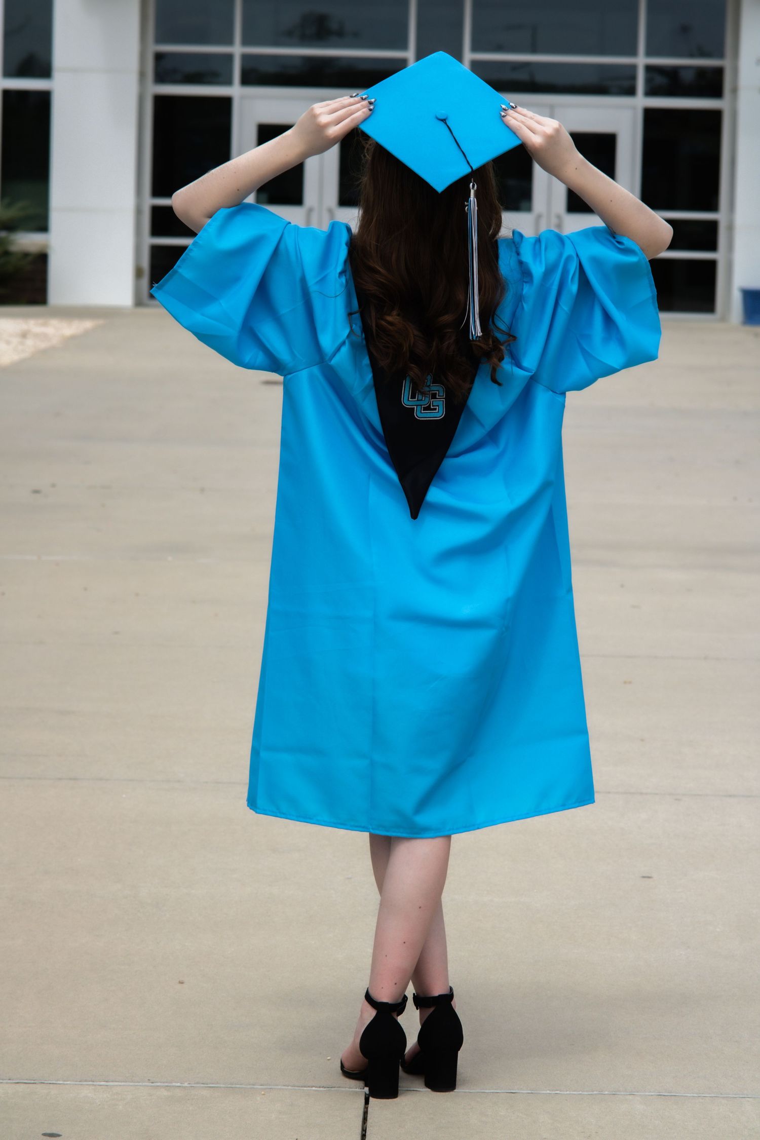 Graduate in bright blue graduation gown adjusts cap while standing outside building.