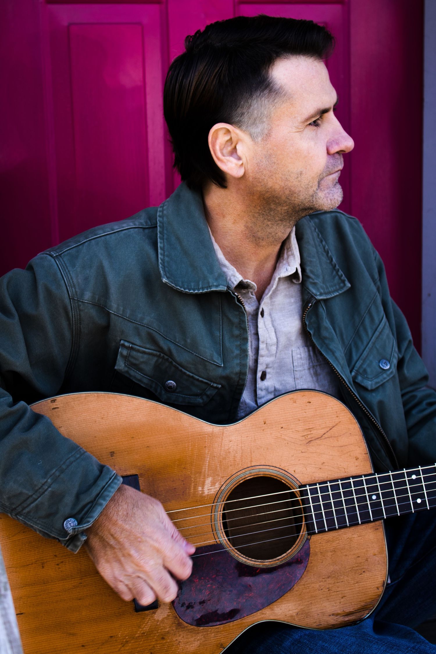 A musician holding an acoustic guitar poses against a bright pink wall.