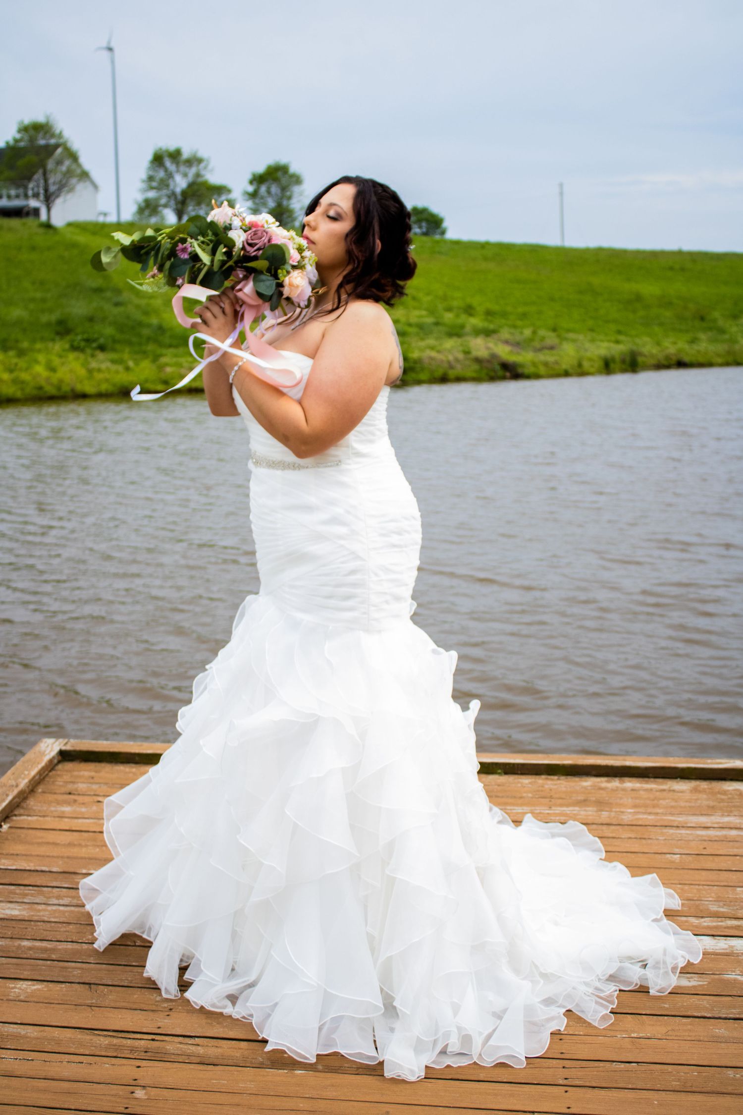 Bride in mermaid wedding dress holding bouquet on dock by lake.