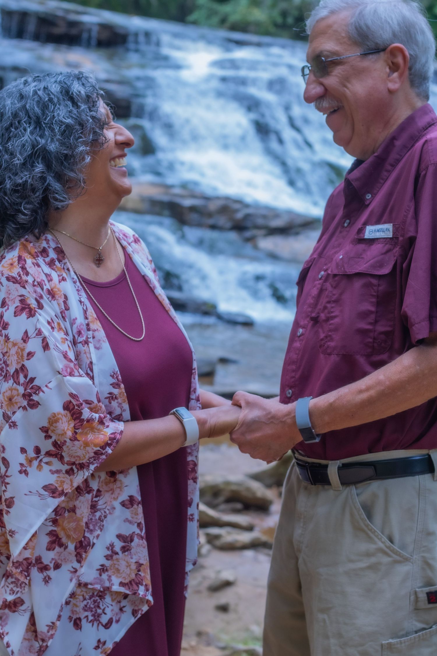 A couple shares a tender moment outdoors near a waterfall, with one wearing burgundy and the other in a floral print.