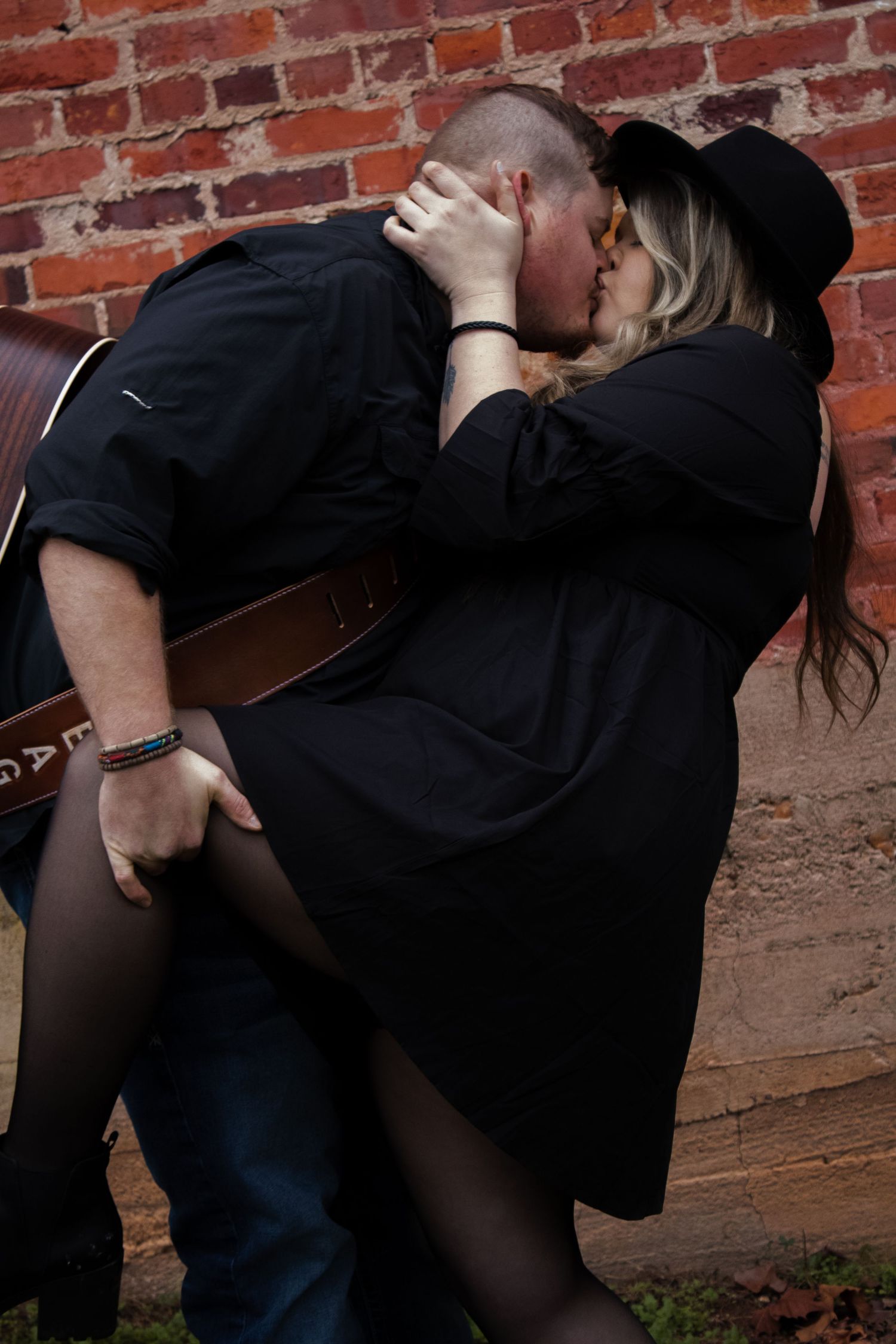 Romantic couple in black clothing embracing against red brick wall.