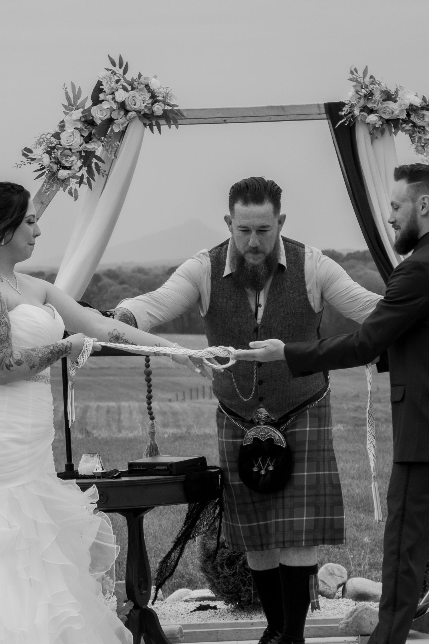 Wedding ceremony under a floral arch with participants in traditional Scottish attire.