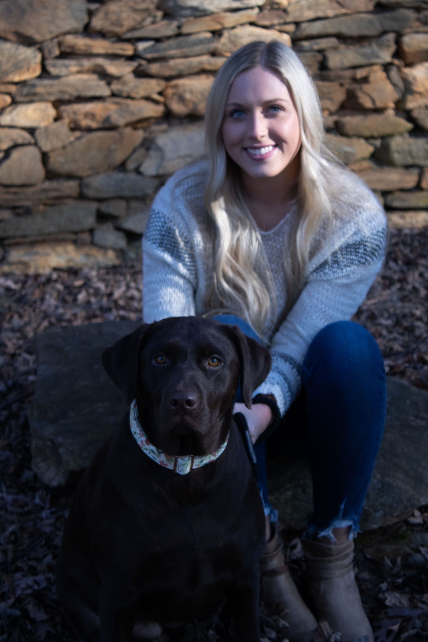 Person in cozy sweater sits with black Labrador dog against rustic stone wall.