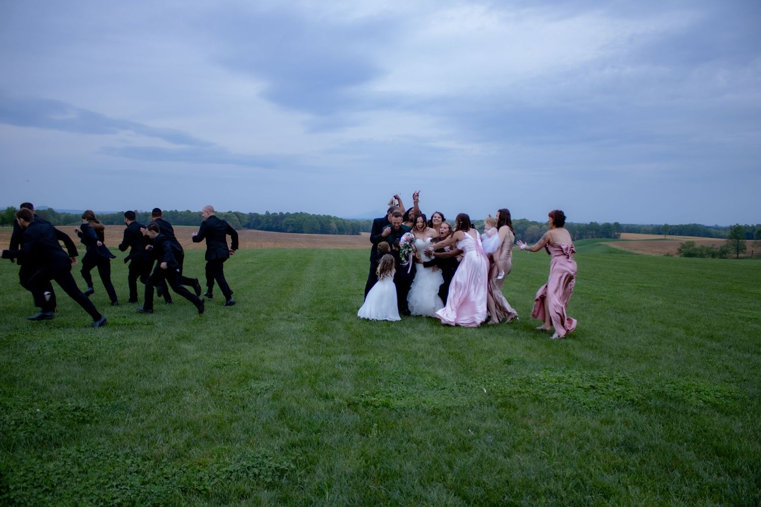 Wedding party running playfully across a green field at twilight.