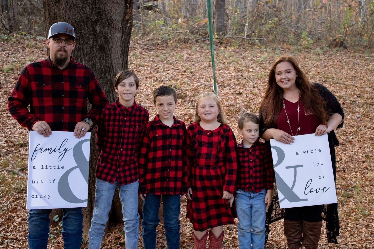 Family portrait with matching buffalo plaid shirts holding decorative signs in fallen leaves.