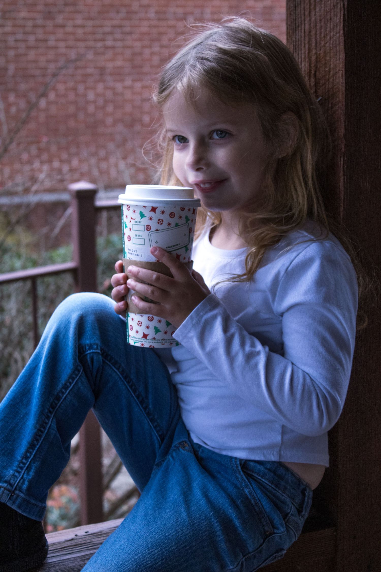 Small child in jeans and white shirt enjoying a drink from a decorated cup while sitting near a brick wall.
