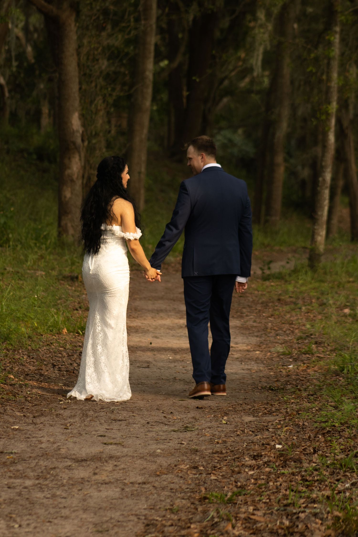 Newlyweds walk hand in hand along a forest path, with bride in white lace gown and groom in navy suit.