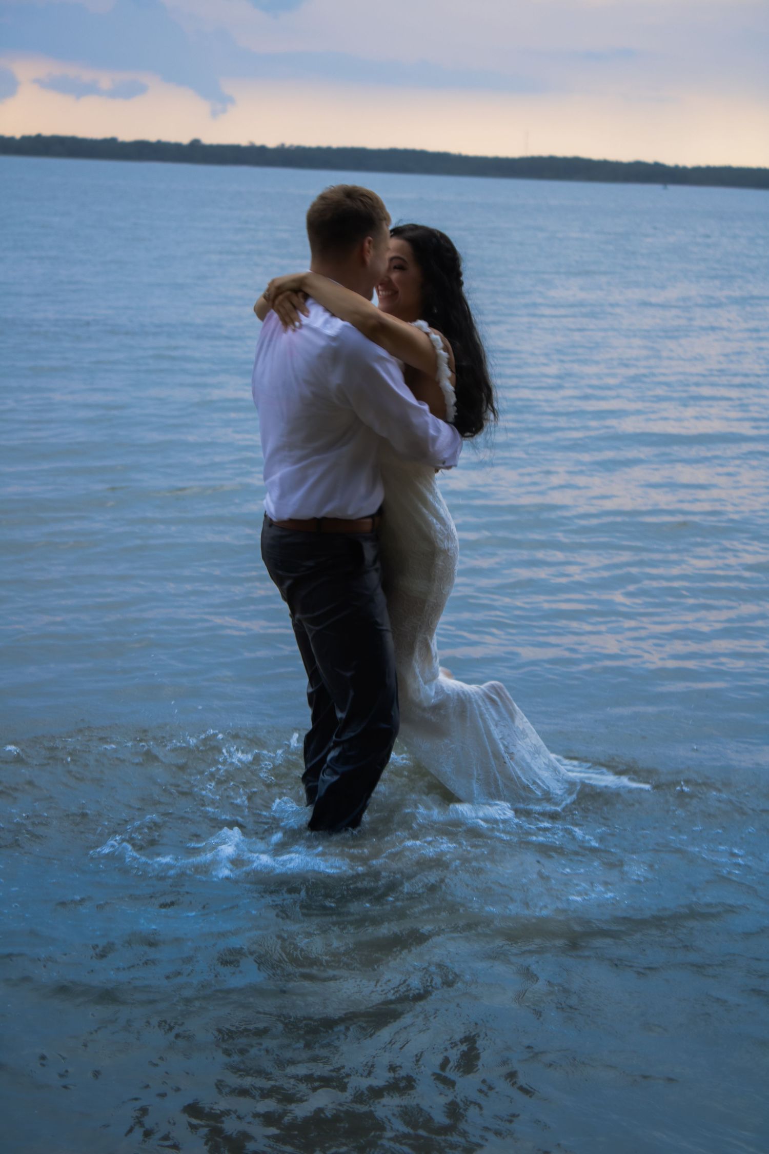 Couple in wedding attire embrace while standing in shallow ocean water at dusk.