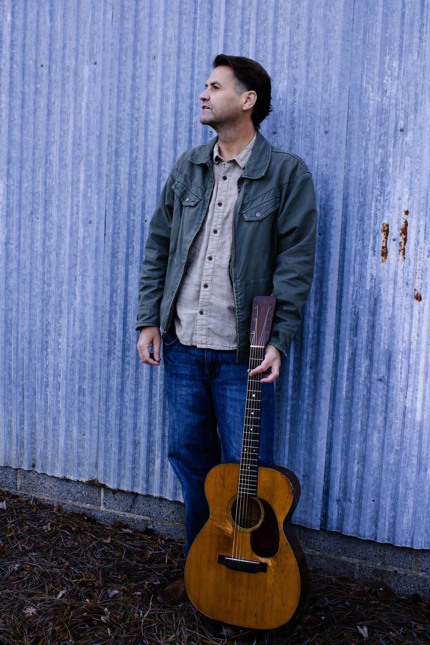 A musician poses with an acoustic guitar against a corrugated metal wall.
