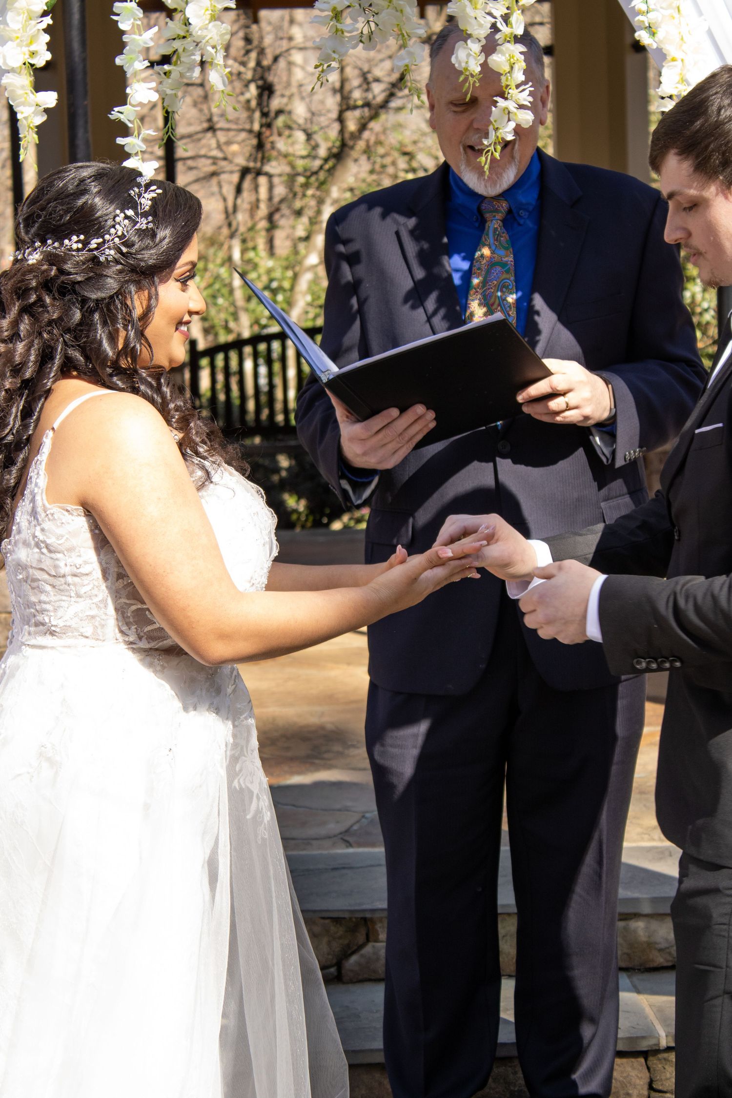 Wedding couple exchanges rings during outdoor ceremony under floral arch.