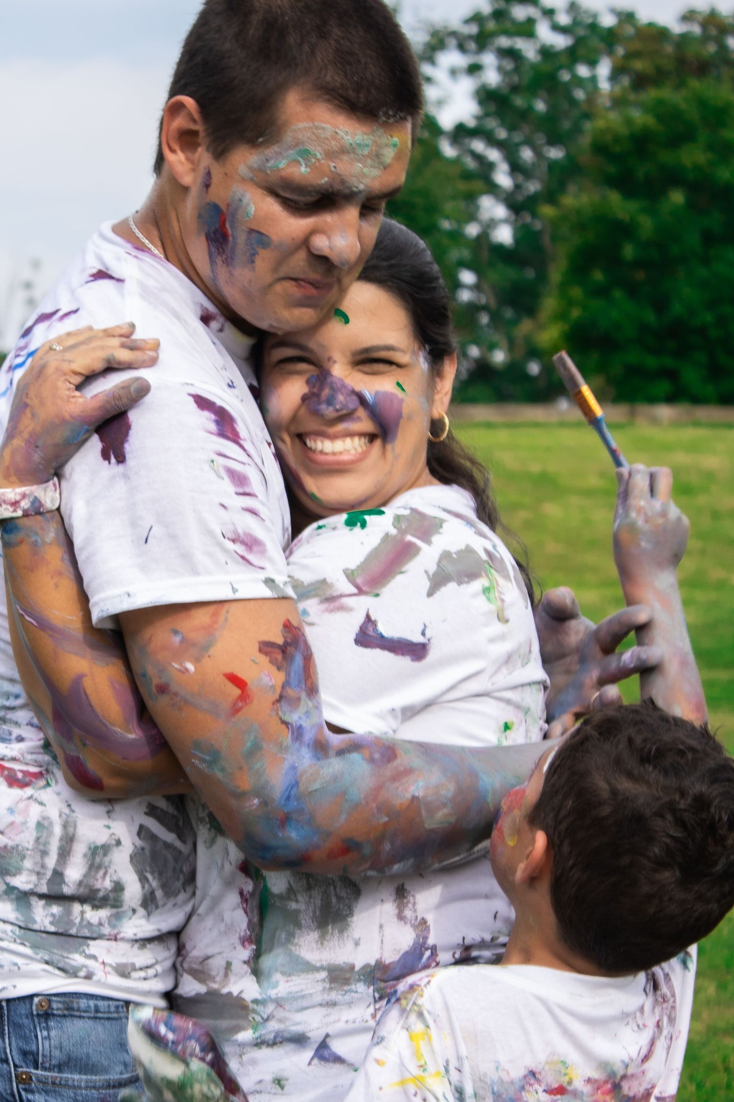 Playful family covered in paint smiles and laughs together on green grass.