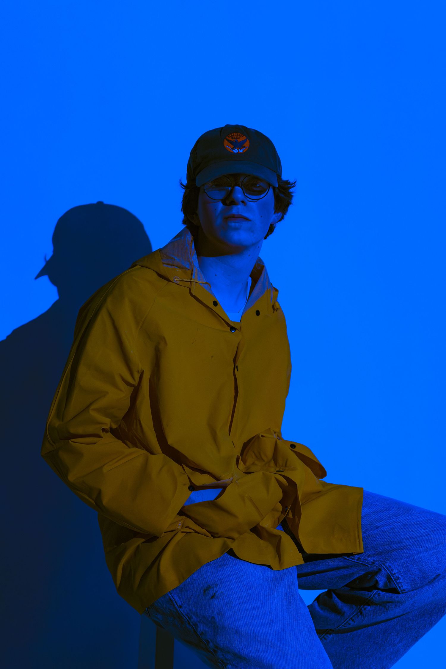 Person in a yellow hoodie and sunglasses sitting against a vivid blue background with dramatic lighting.