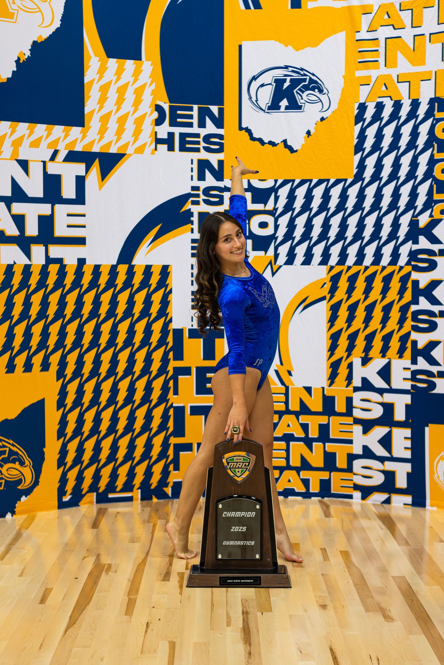 Three cheerleaders in blue uniforms perform in front of a yellow and blue patterned wall in a gym.