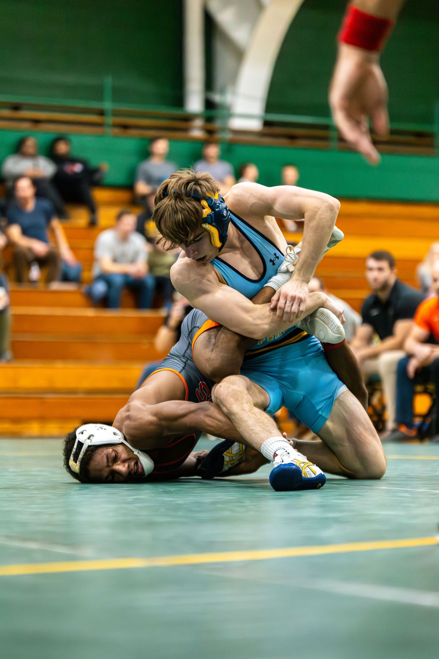 Two wrestlers compete on a green mat during an intense match with spectators visible in the background.