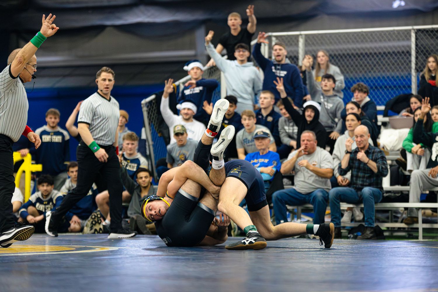 Wrestling match with athletes competing on mat while spectators cheer excitedly from bleachers in background.