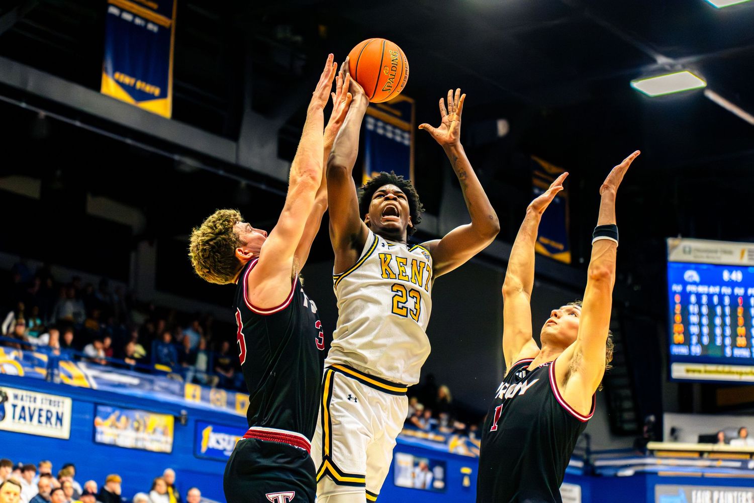 Basketball player in gold jersey attempts a shot while being defended by two players in black jerseys during game action.