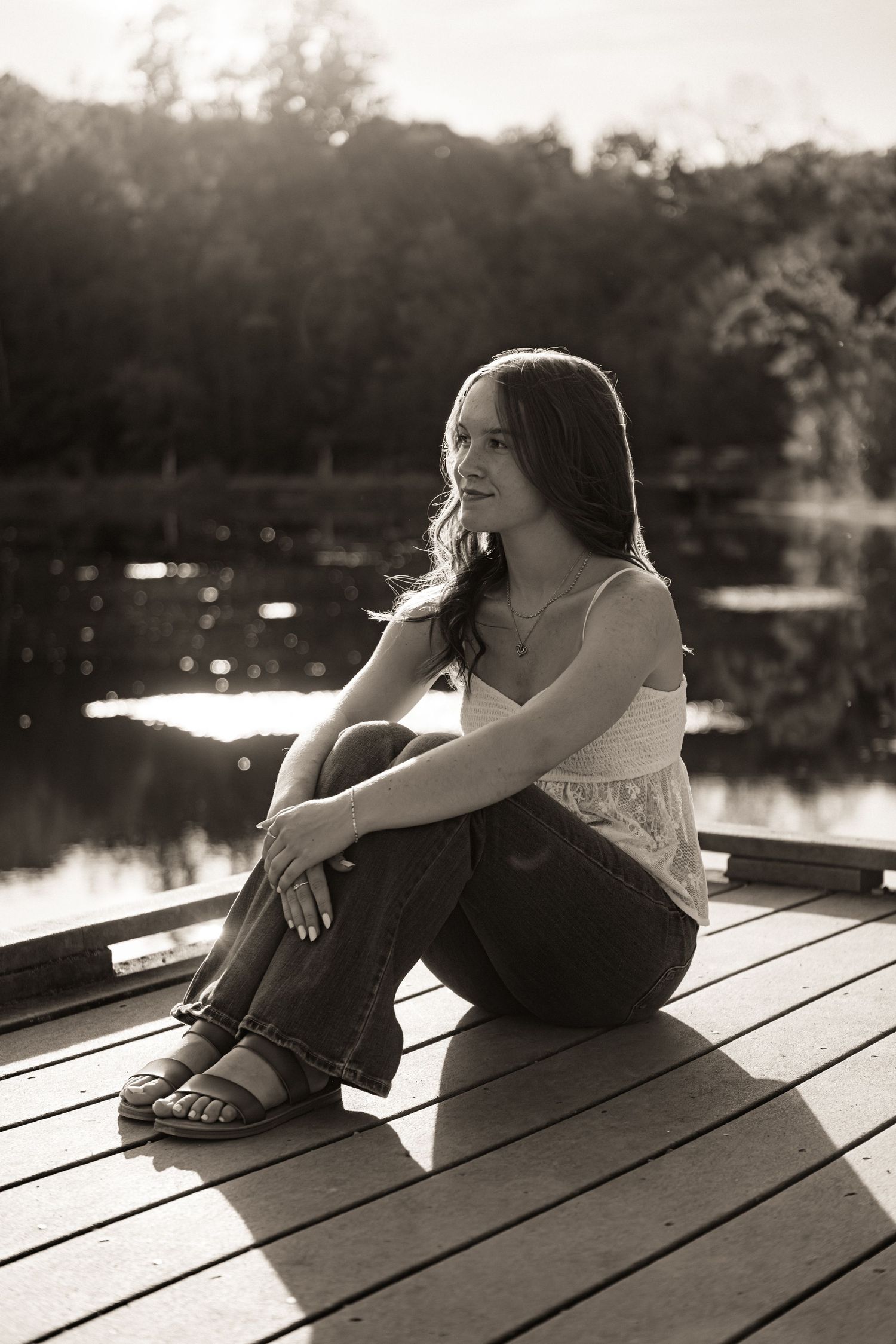 A person sits thoughtfully on a wooden dock by a lake during sunset in a black and white photograph.