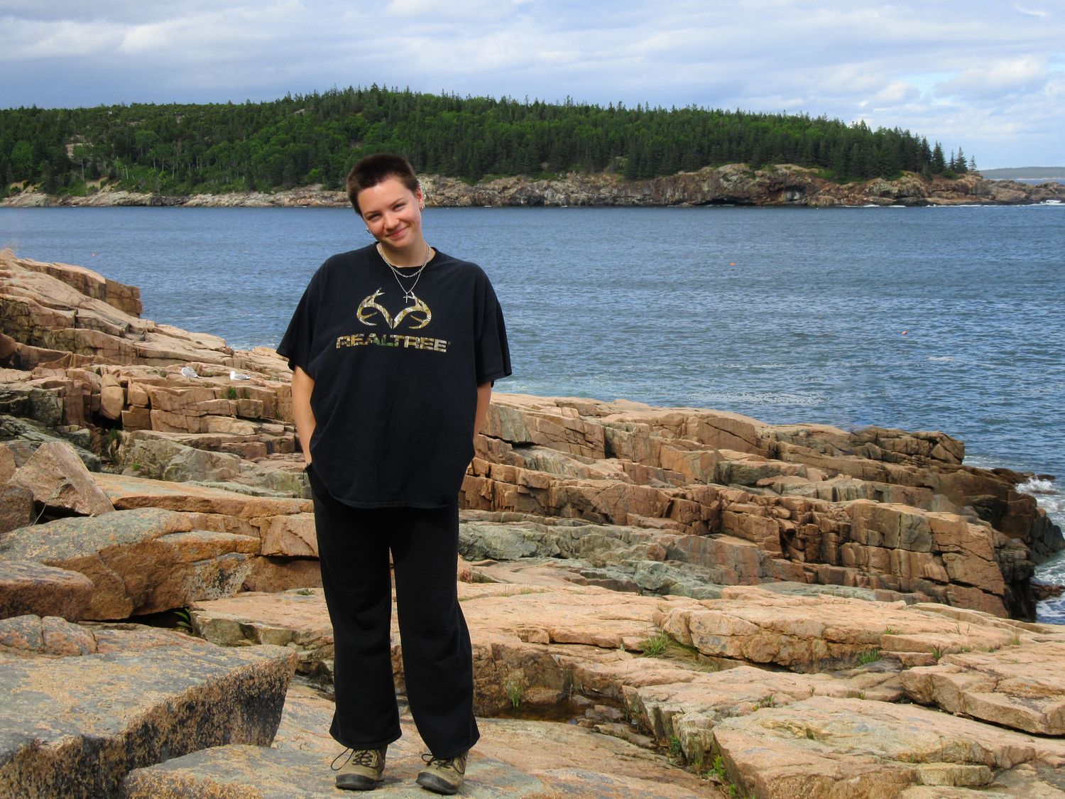 Person standing on rocky coastal shore wearing a Realtree shirt, with blue ocean waters and forested hills behind.