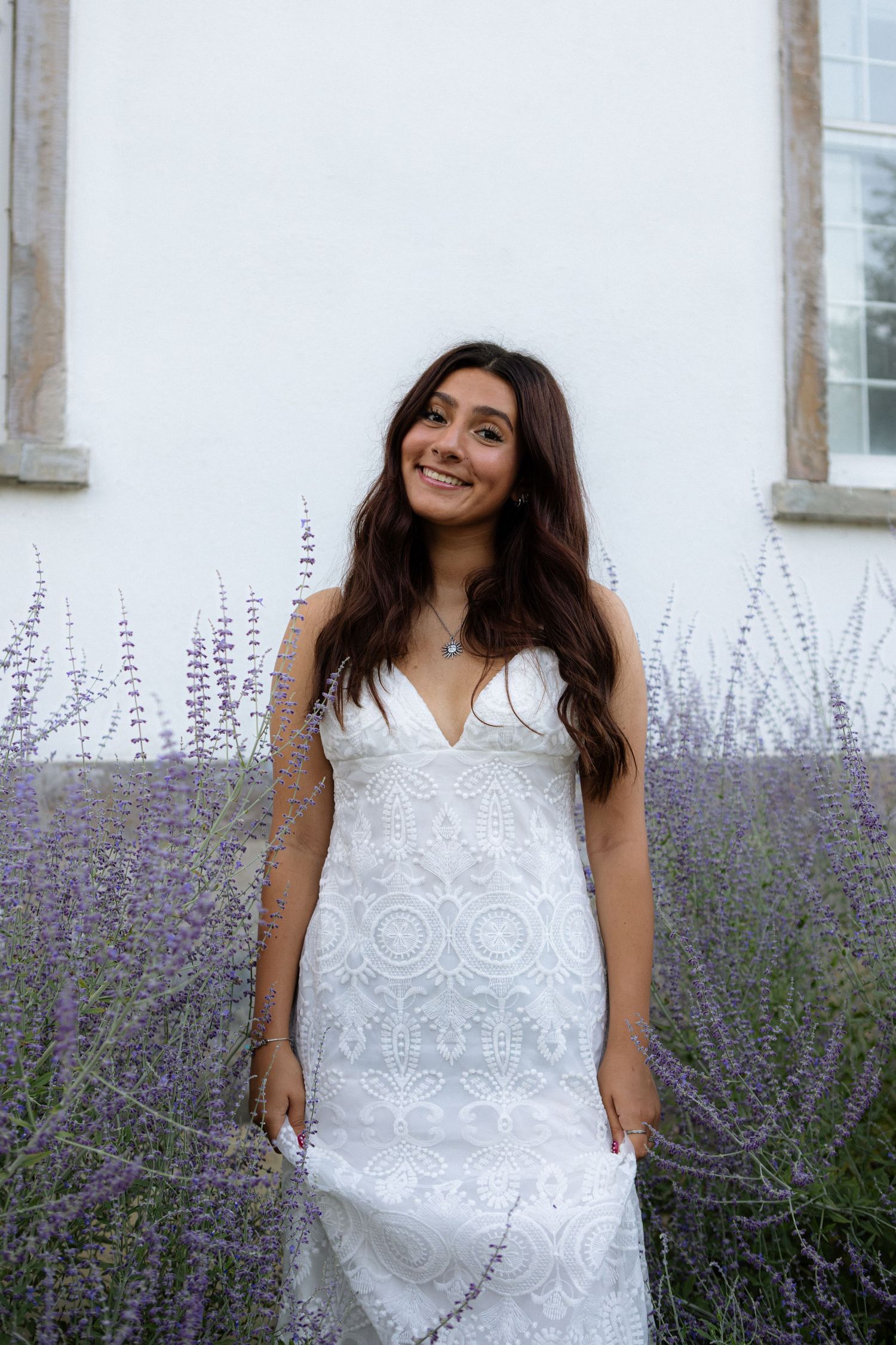 A white lace dress worn in front of blooming lavender plants and a white wall.